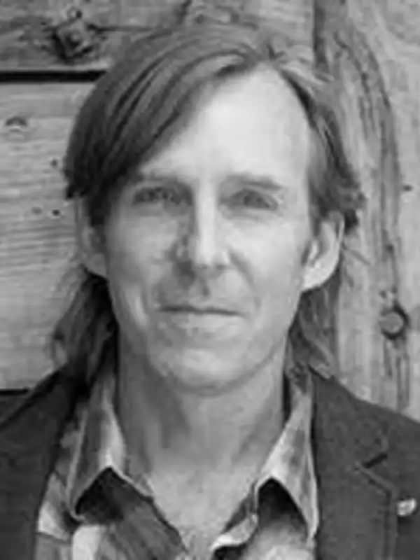 A white man with long light hair smiles into the camera in black and white.